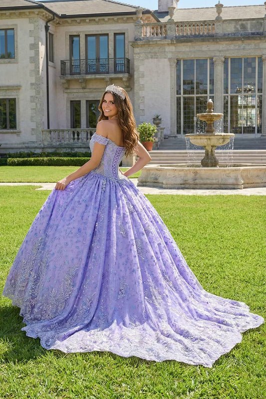 Woman in a lavender ball gown with floral embroidery and tiara poses outdoors by a fountain